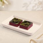 Radhe Cranberry Exotica, two squares of red cranberry katri with a dark chocolate top and pistachio decoration on a white tray
